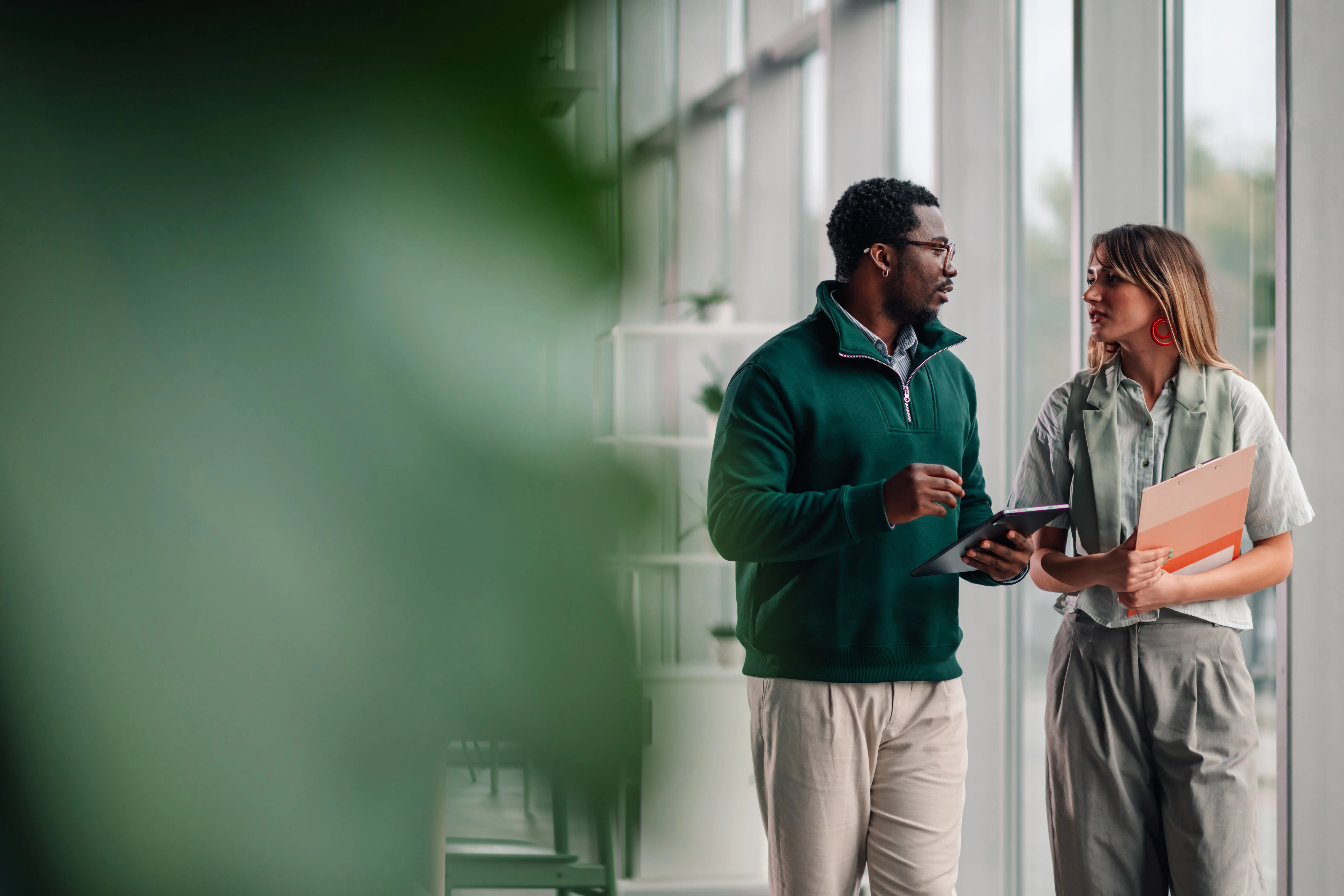 Two colleagues having a discussion in a modern office corridor; the man holds a tablet and wears glasses with a green pullover, while the woman holds a clipboard with documents and wears gray pants and a light shirt.