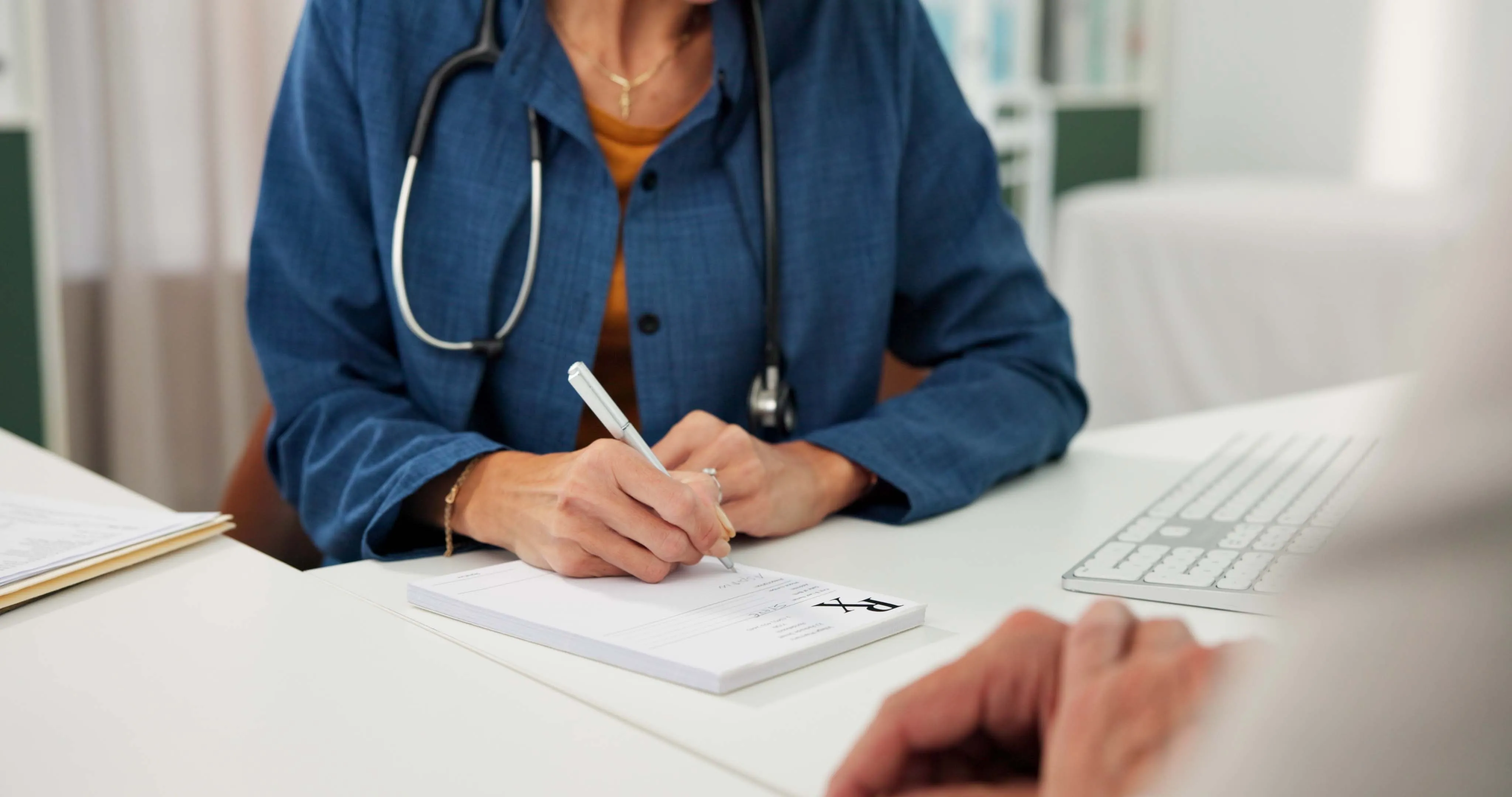 Medical professional wearing a blue coat and stethoscope writes a prescription on a notepad marked with RX, sitting at a desk with a keyboard nearby and a patient's hand visible in foreground.