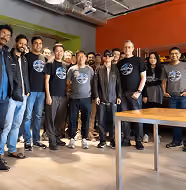 Group of diverse people standing indoors in a casual workspace with bright orange and green walls, some wearing matching t-shirts with a circular logo.