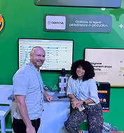 Two smiling people interacting at a technical workspace with a computer screen and equipment, set against a green background with text about operating a production machine.