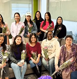 A diverse group of eleven women smiling and holding wrapped roses, gathered indoors in front of a plain white wall and windows.