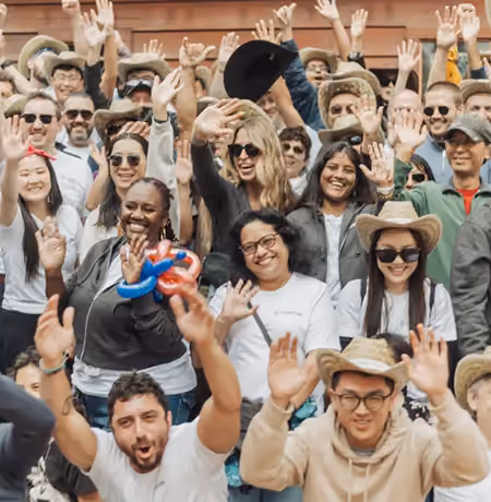 Diverse group of adults smiling and waving at the camera, some wearing sunglasses and hats, gathered outdoors in front of a wooden background.