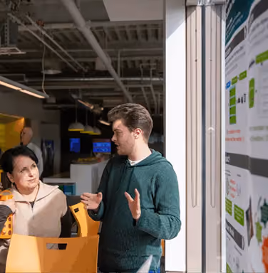 Two people having a discussion indoors: a woman with black hair wearing a beige jacket holding a yellow object, and a man with brown hair in a green sweater gesturing with his hands, with office and information boards in the background.