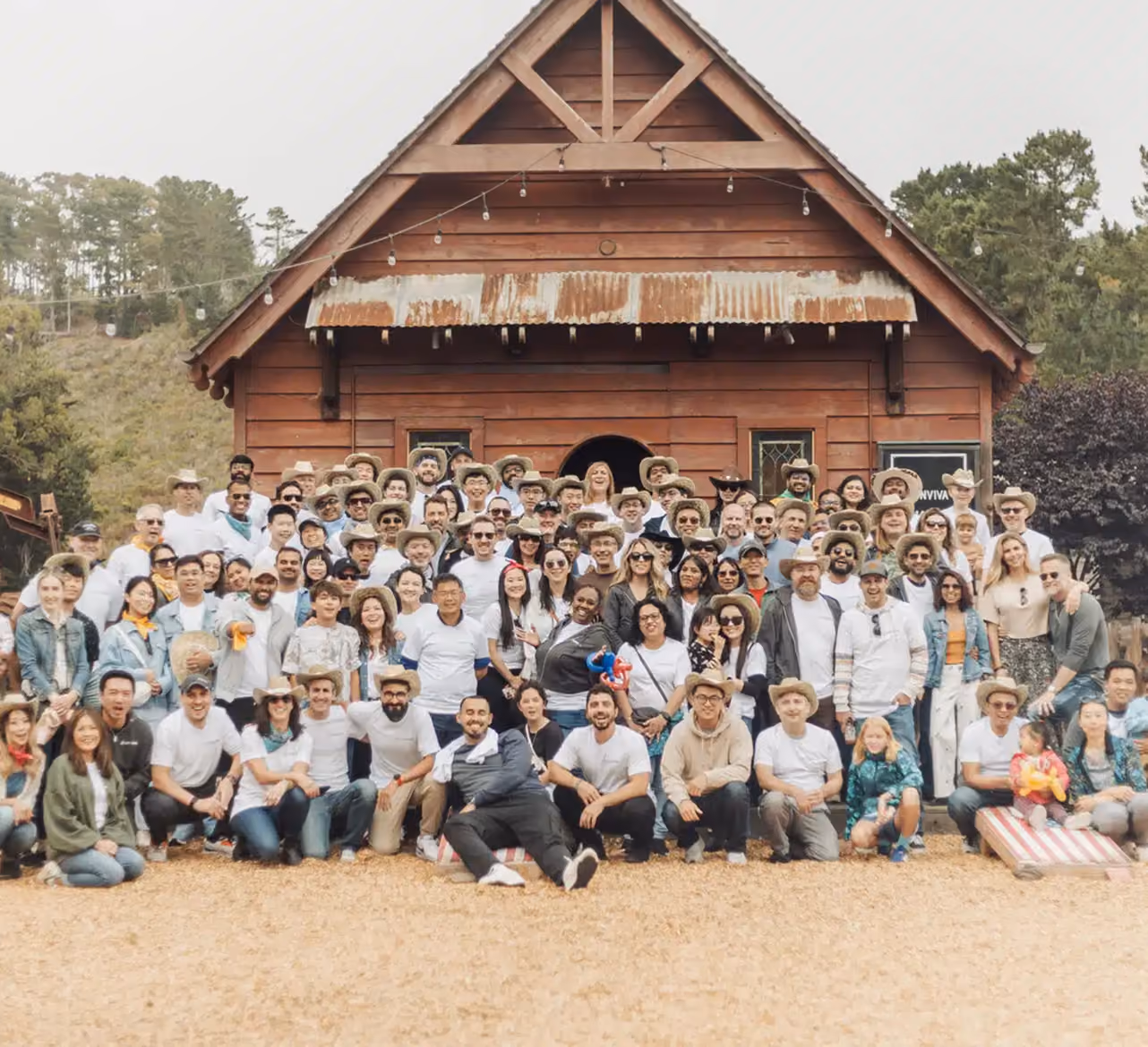 Large group of people posing for a photo outdoors in front of a rustic wooden cabin with a corrugated metal awning, many wearing cowboy hats and casual clothing, on a gravel or dirt ground with trees in the background.