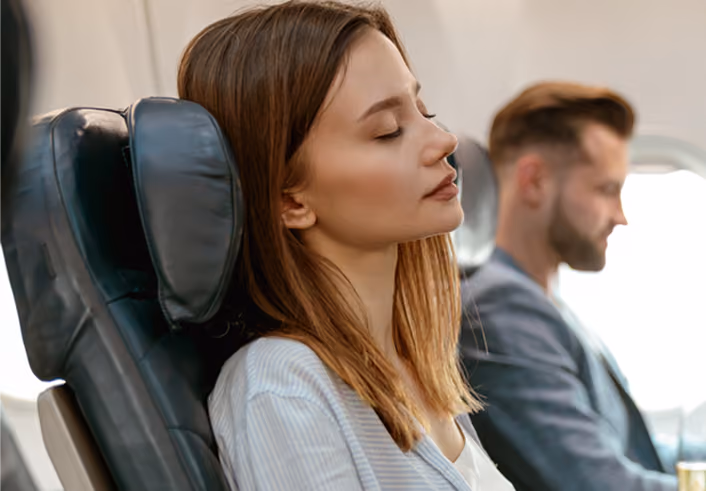 Woman with brown hair resting with eyes closed in an airplane seat with a headrest, man sitting in background.