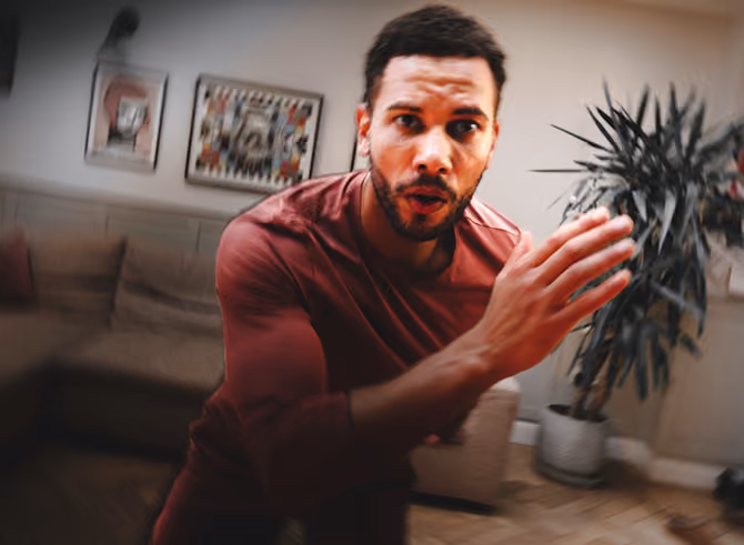 Man in a red long-sleeve shirt reaching forward with an intense expression in a living room with a sofa, framed pictures, and a potted plant.