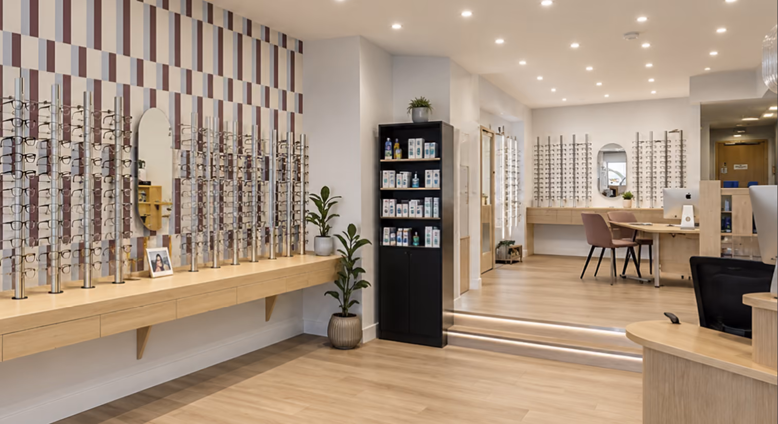 Modern optician store interior with wooden floors and light walls featuring multiple vertical racks displaying eyeglass frames, two oval mirrors, potted plants, a black shelving unit with eye care products, and desks with chairs and computers.