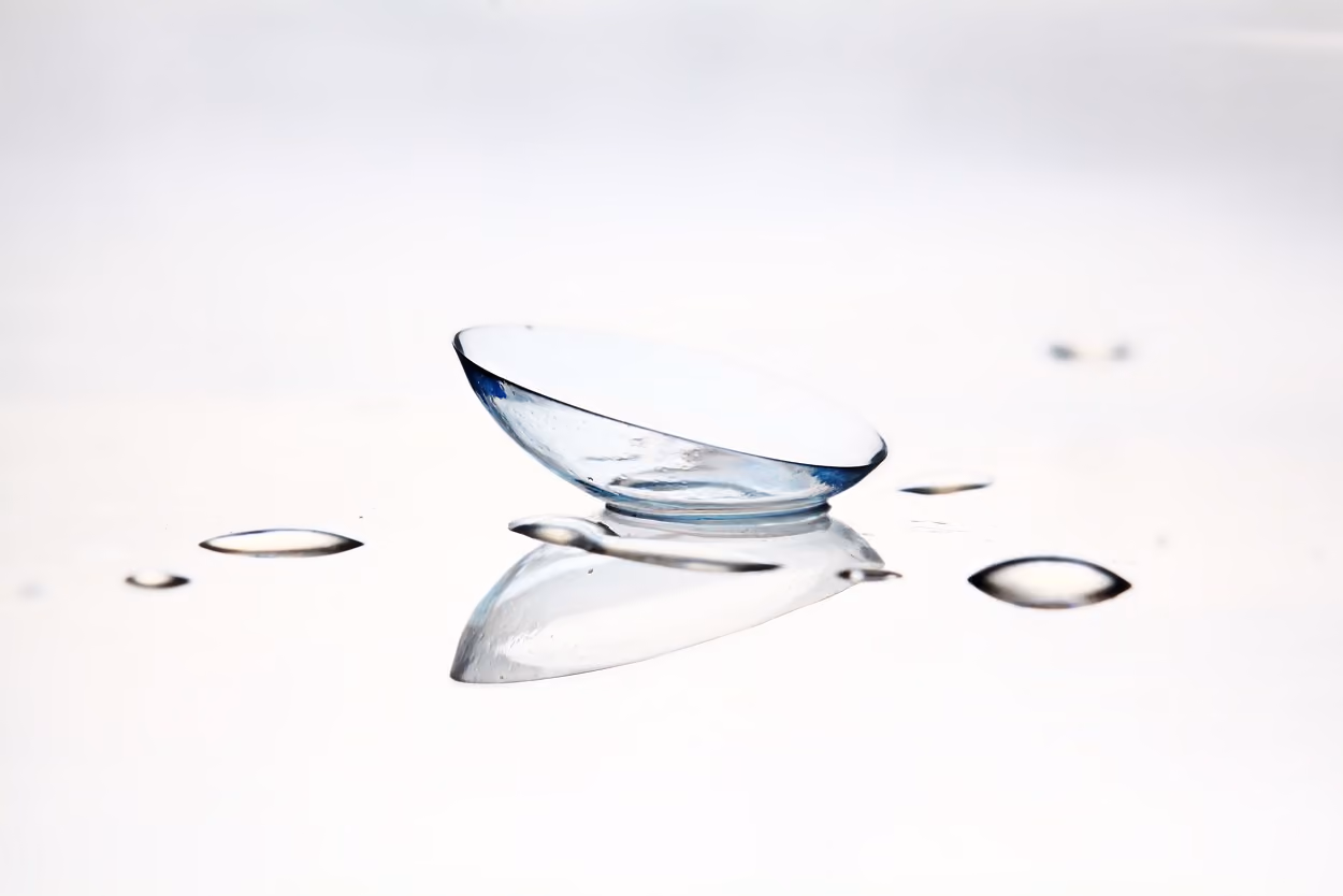 A single transparent contact lens resting on a smooth white surface with small water droplets surrounding it.