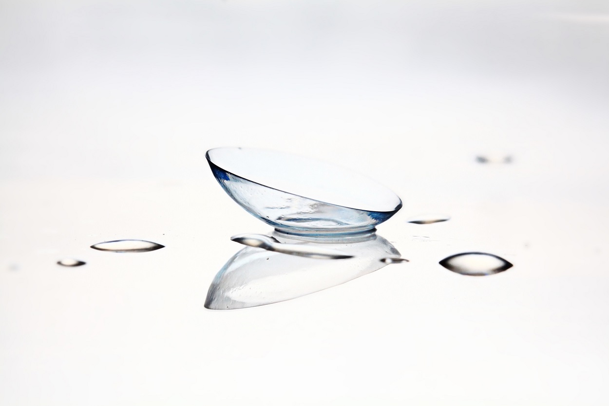 A single transparent contact lens resting on a smooth white surface with small water droplets surrounding it.