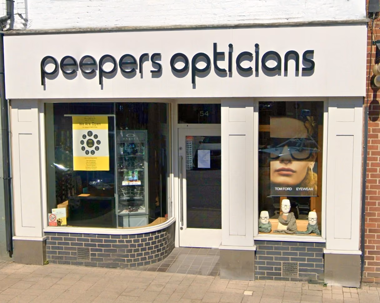 Front view of Peepers Opticians store with white facade and large black lettering, two display windows showcasing eyewear including a poster of a woman wearing sunglasses and mannequin heads with glasses.