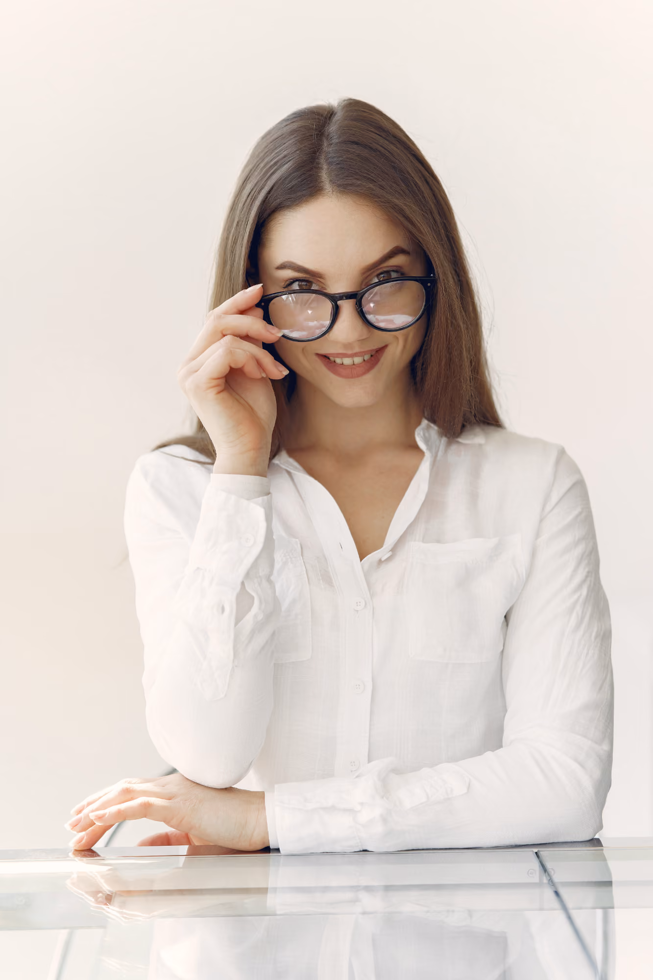 A woman with long brown hair wearing a white button-up shirt is smiling and looking over the top of her black-framed glasses while resting her arms on a reflective glass surface.