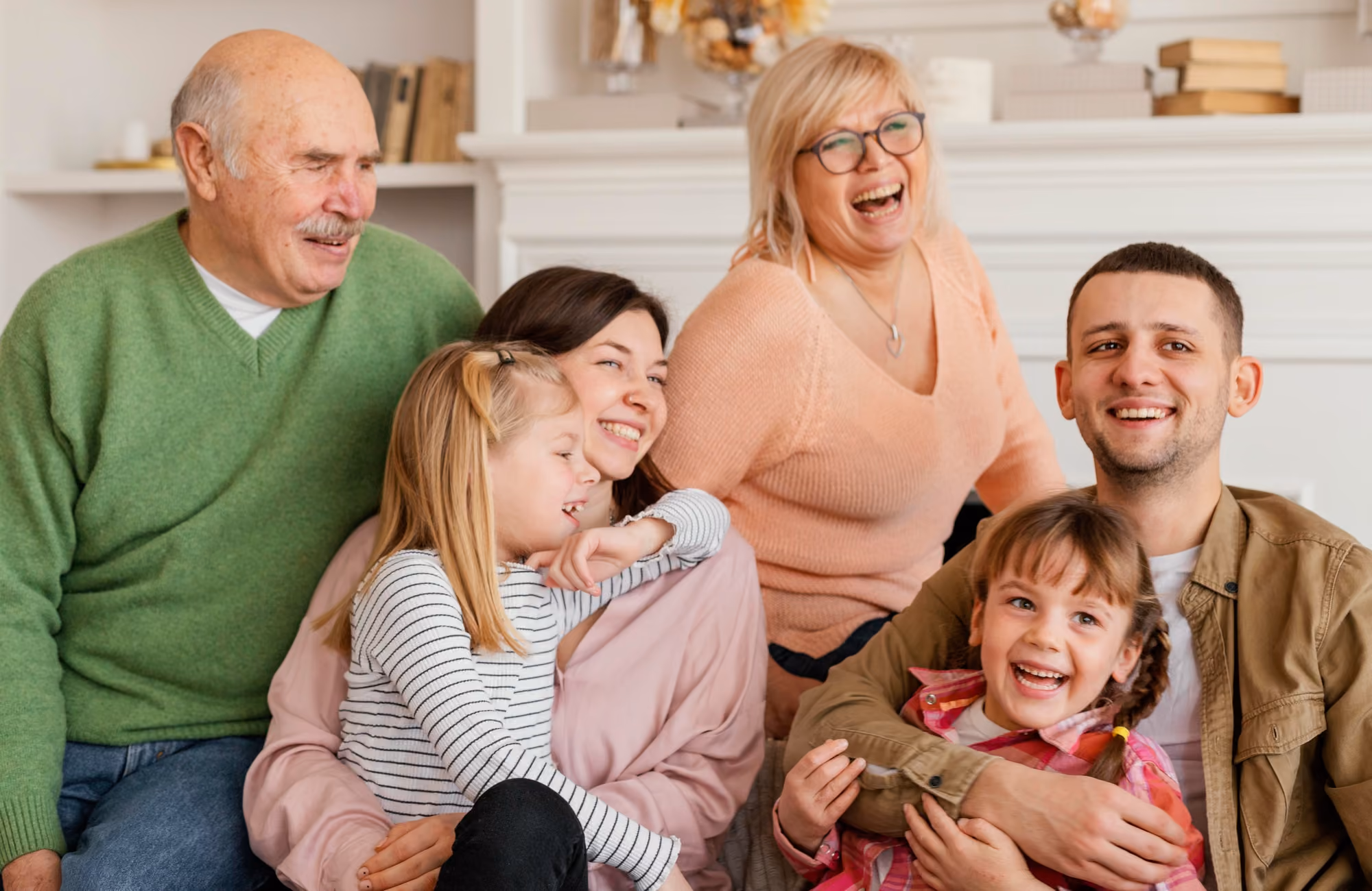 A joyful multigenerational family sitting together in a cozy living room, with a grandfather in a green sweater, a grandmother in a peach sweater laughing, a mother holding a blonde girl in a striped shirt, and a father hugging a smiling girl in a pink plaid shirt.