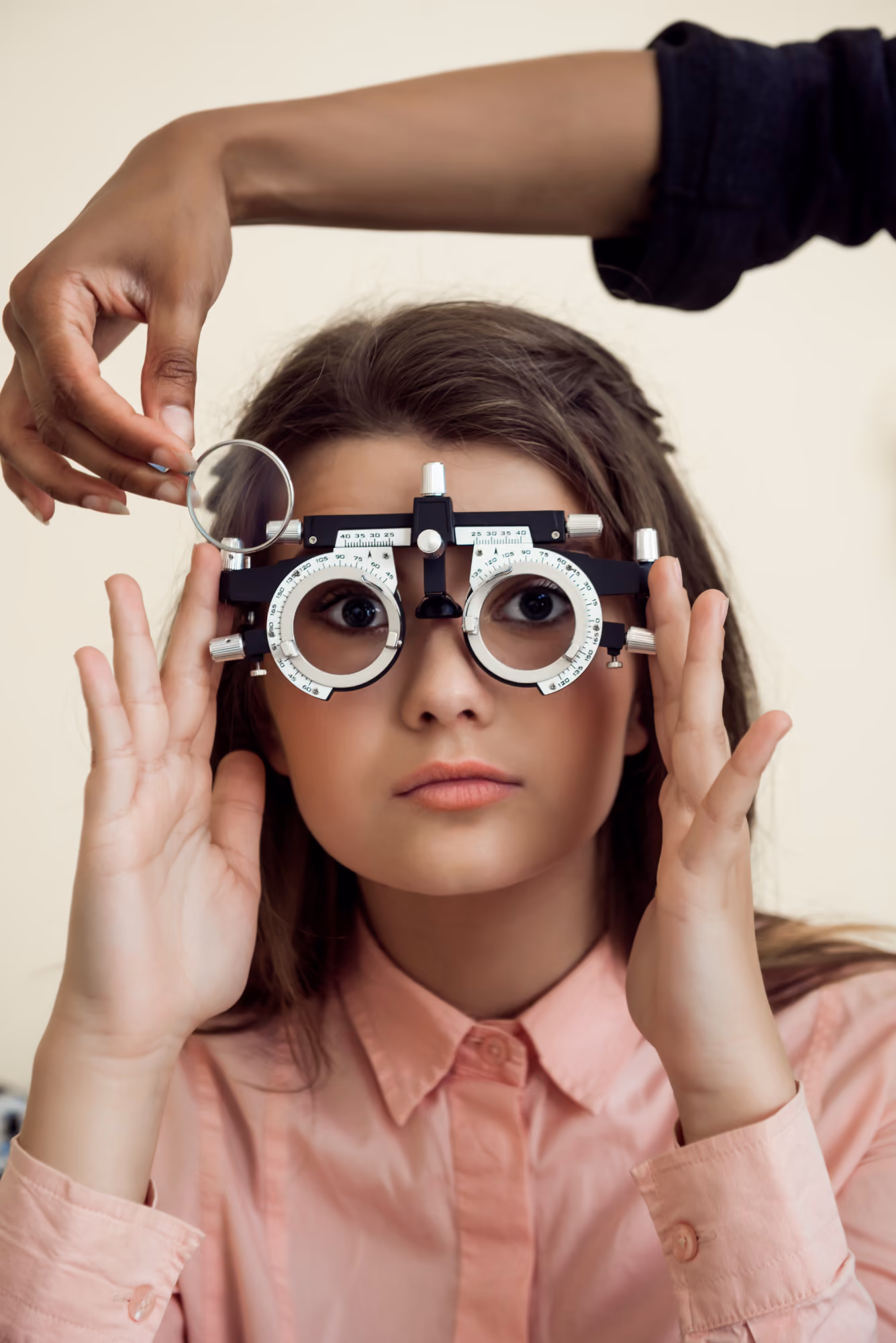Young woman wearing a pink shirt is undergoing an eye test, holding an adjustable optometry device with round lenses, while a second person adjusts a lens on the device.