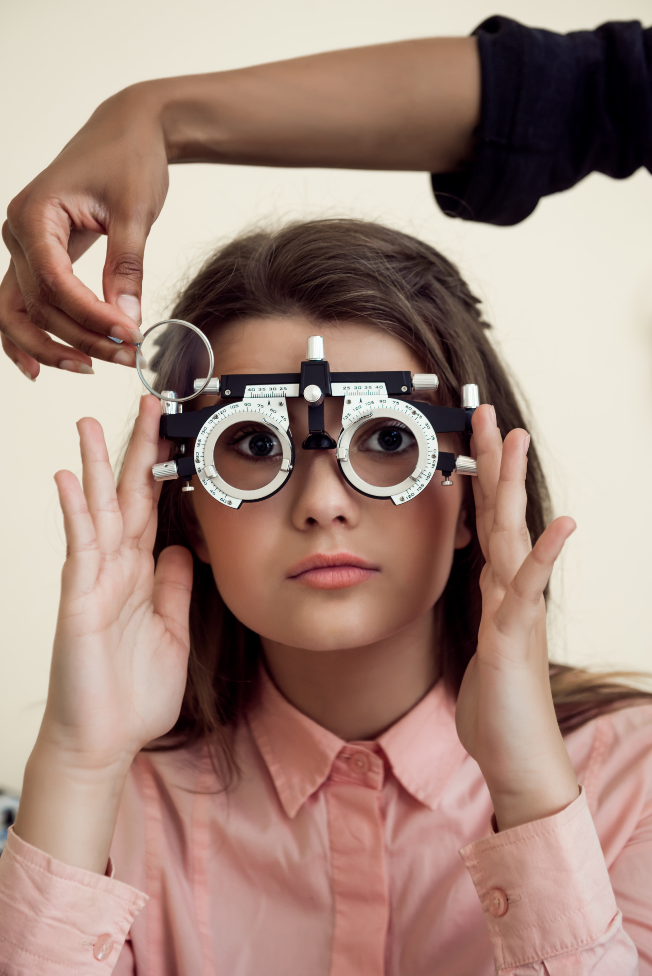 Young woman wearing a pink shirt is undergoing an eye test, holding an adjustable optometry device with round lenses, while a second person adjusts a lens on the device.