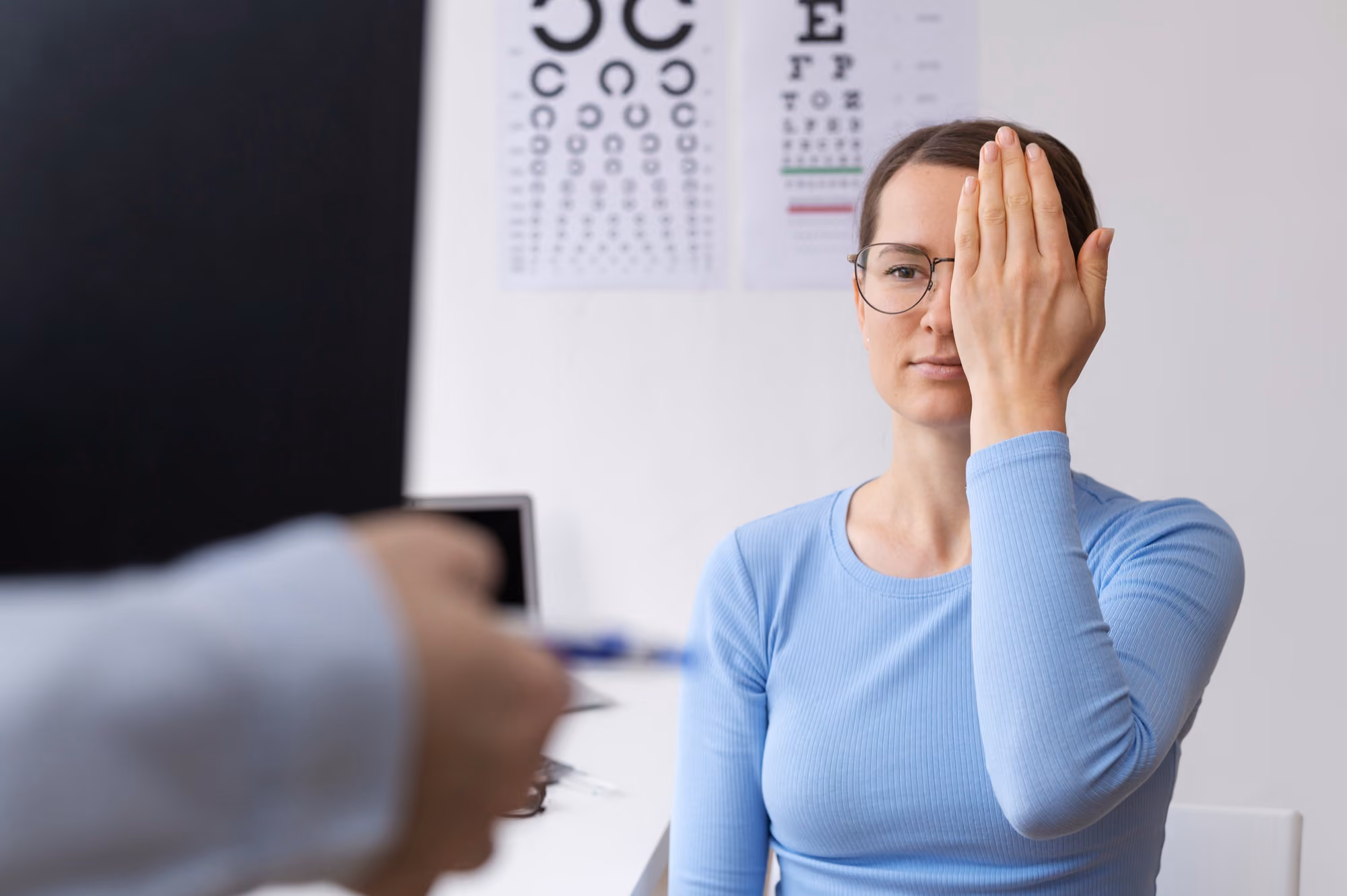 Woman wearing glasses covers one eye with her hand while undergoing an eye exam with a vision test chart in the background.