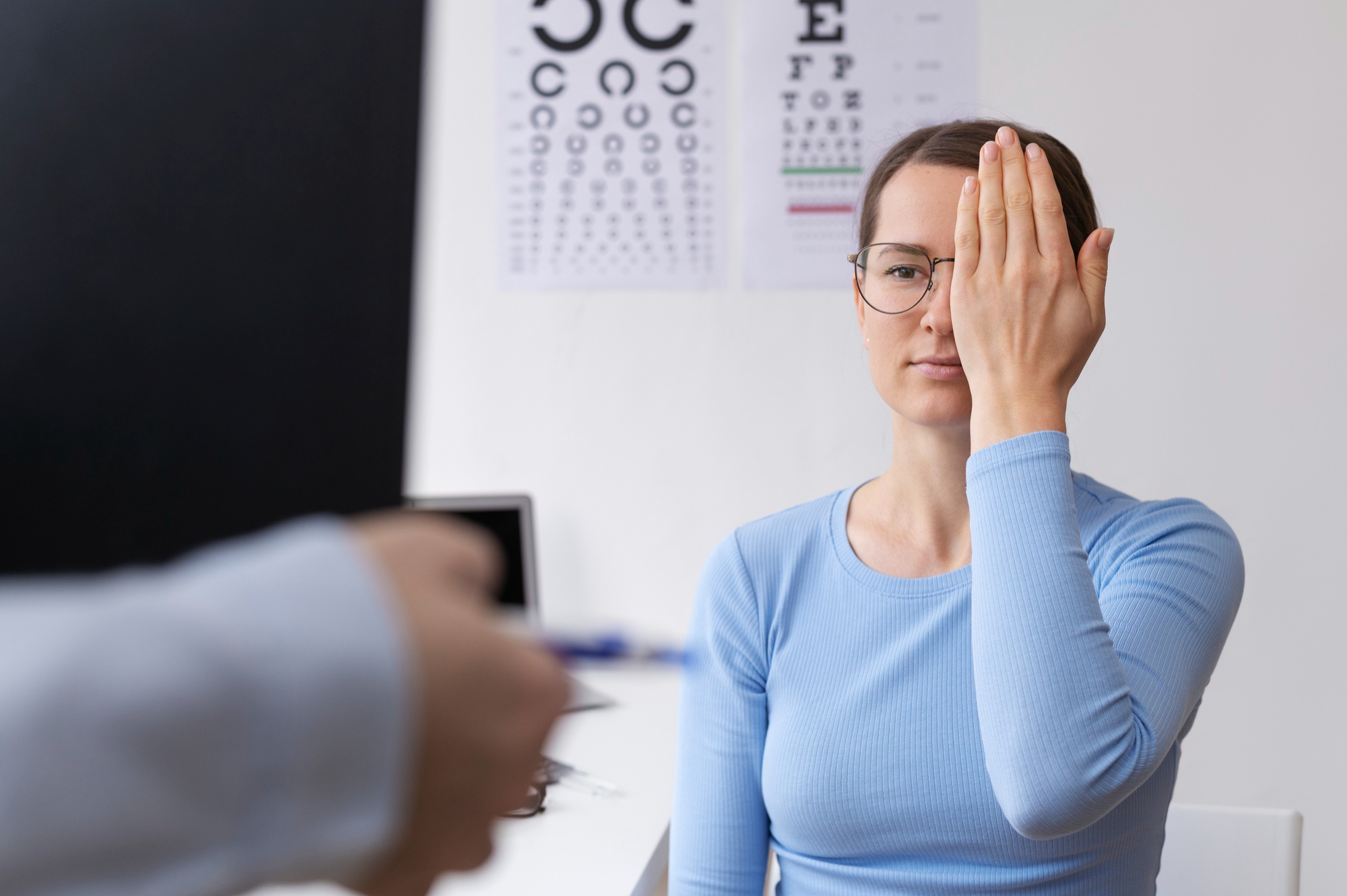 Woman wearing glasses covers one eye with her hand while undergoing an eye exam with a vision test chart in the background.
