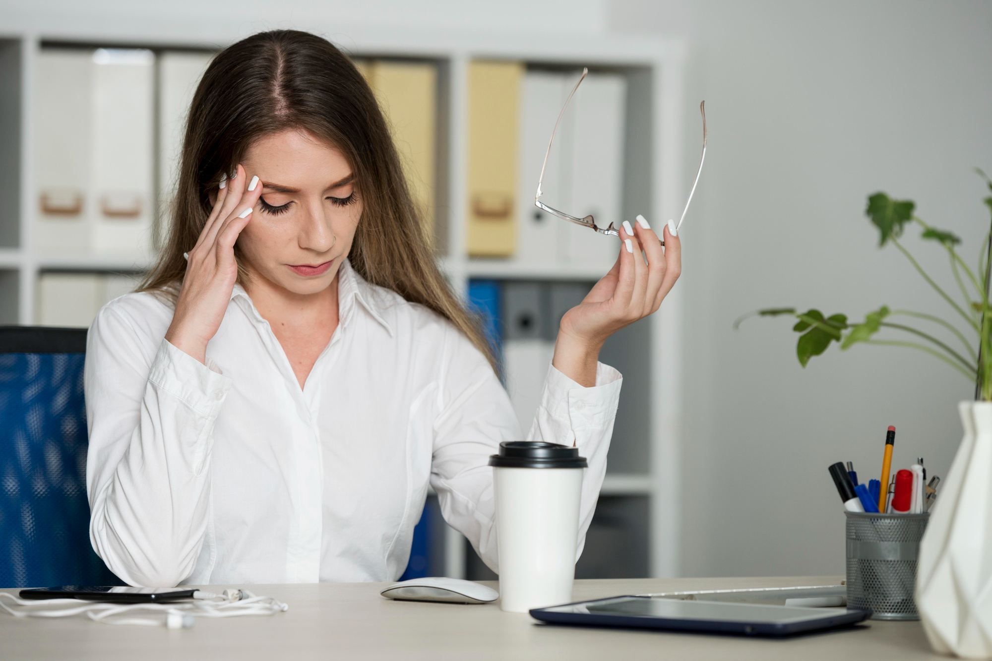 A woman in a white blouse sits at a desk holding her glasses in one hand and touching her temple with the other, appearing stressed or tired, with office supplies and a coffee cup on the desk.