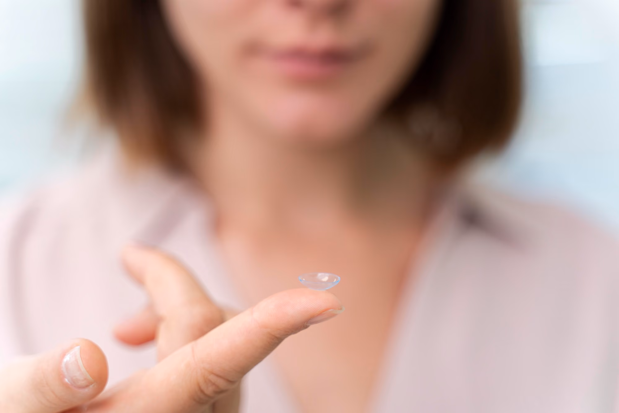 A person holding a clear contact lens on the tip of their index finger, with a blurred face and upper body in the background.