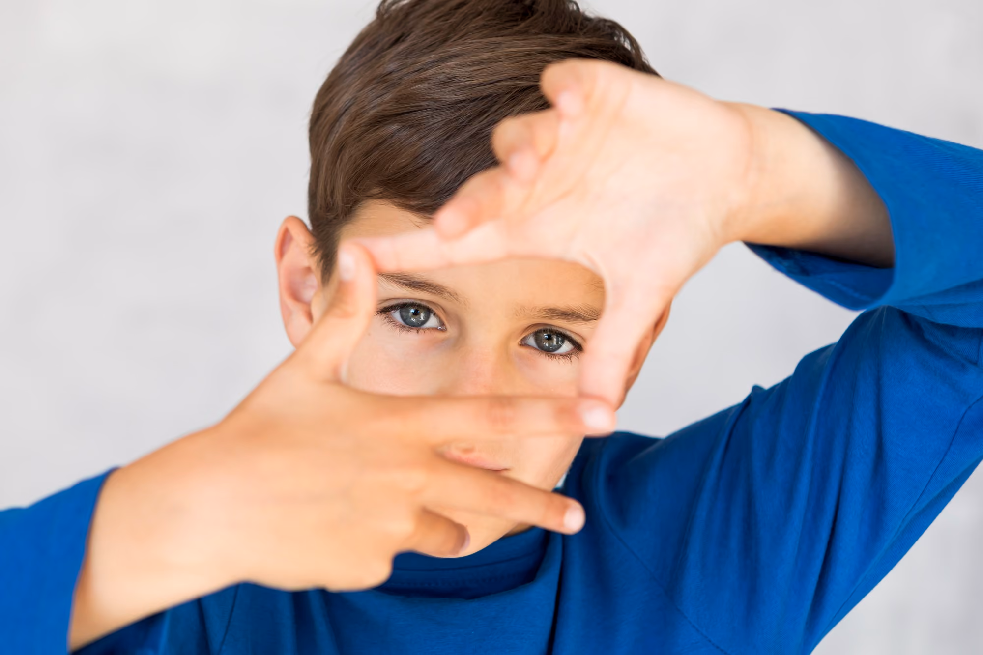 Close-up of a young boy wearing a blue long-sleeve shirt framing his face with his fingers and looking through the frame at the camera with blue eyes.