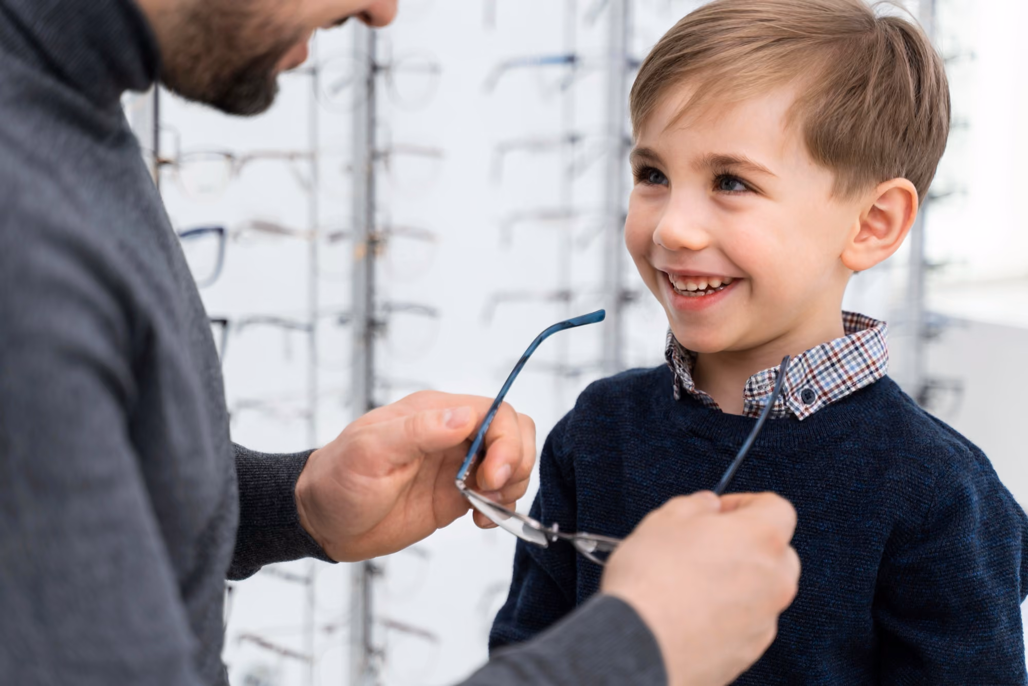 A smiling young boy in a navy sweater and plaid-collar shirt looks at an adult holding eyeglasses in an optical store with glasses displayed in the background.