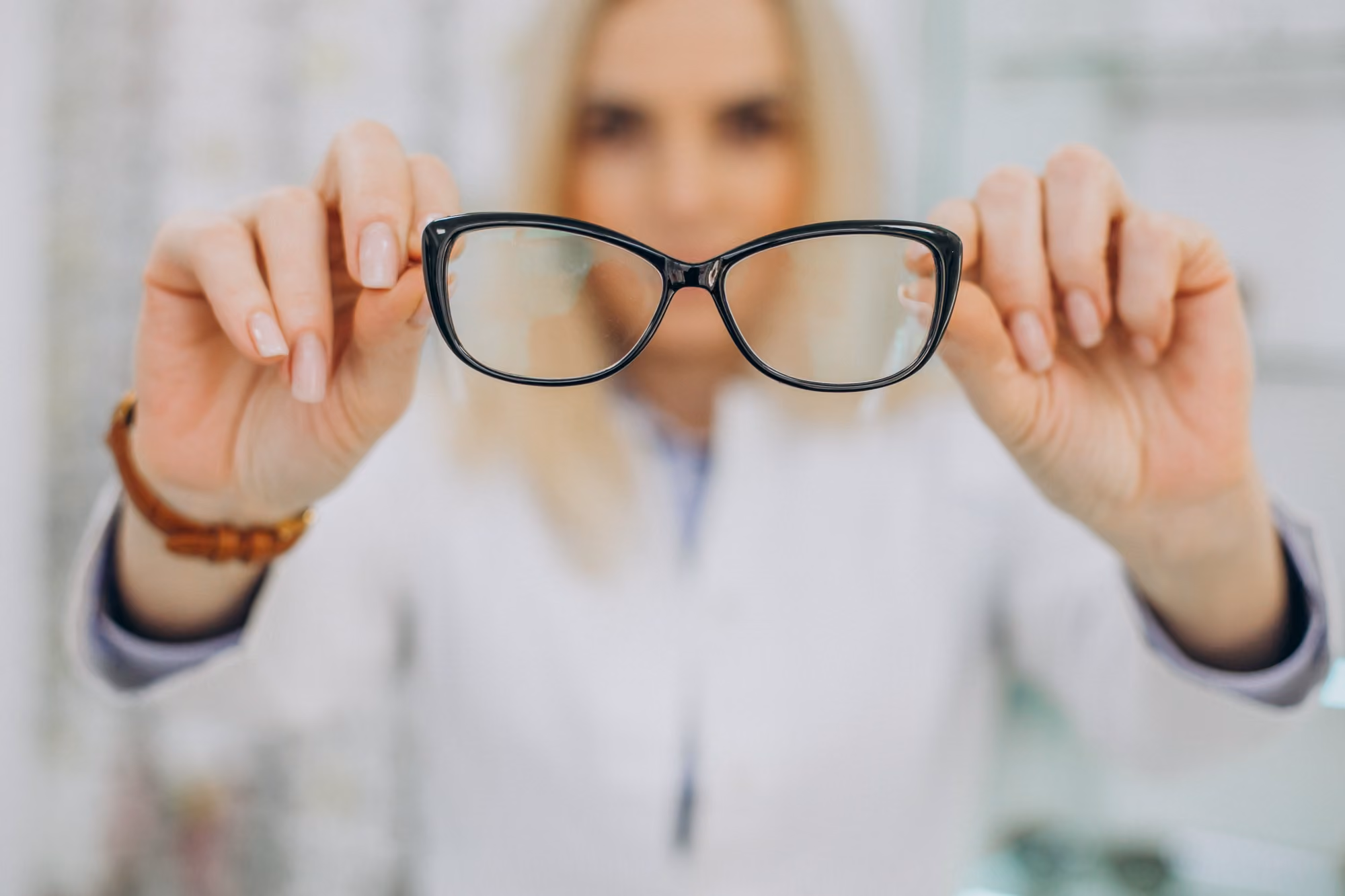 Close-up of black eyeglass frames held by a person wearing a white coat, with the person blurred in the background.