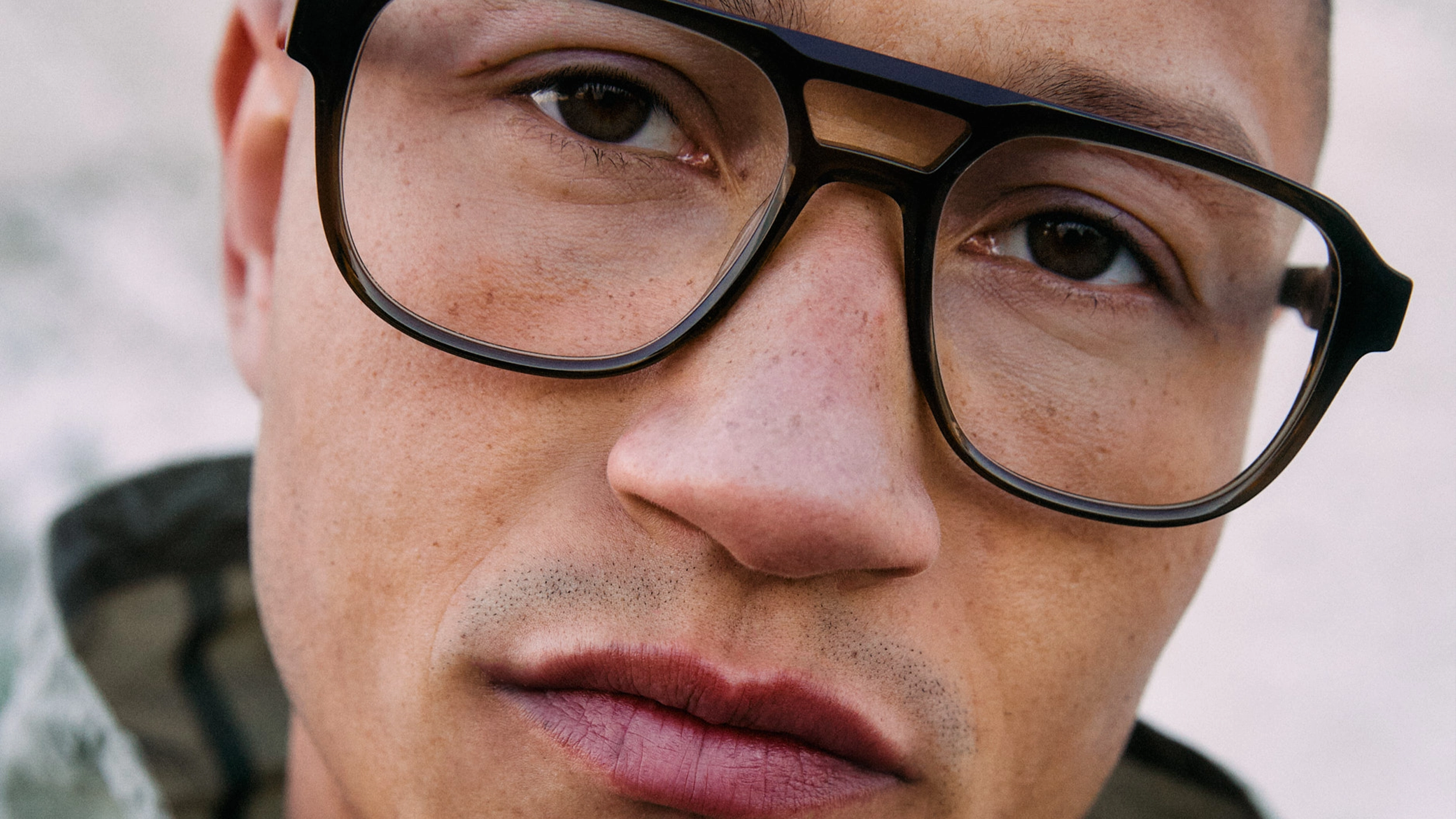 Close-up of a man's face wearing large, thick-rimmed glasses with brown eyes, slightly pursed lips, and visible skin texture including freckles and facial hair stubble.