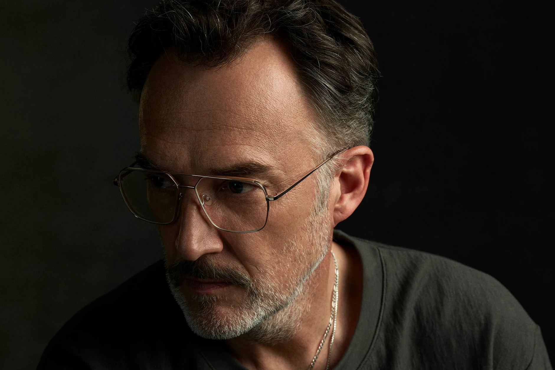 Close-up portrait of a middle-aged man with light stubble and mustache wearing large, thin-framed glasses and a dark shirt, looking thoughtfully to the side against a dark background.