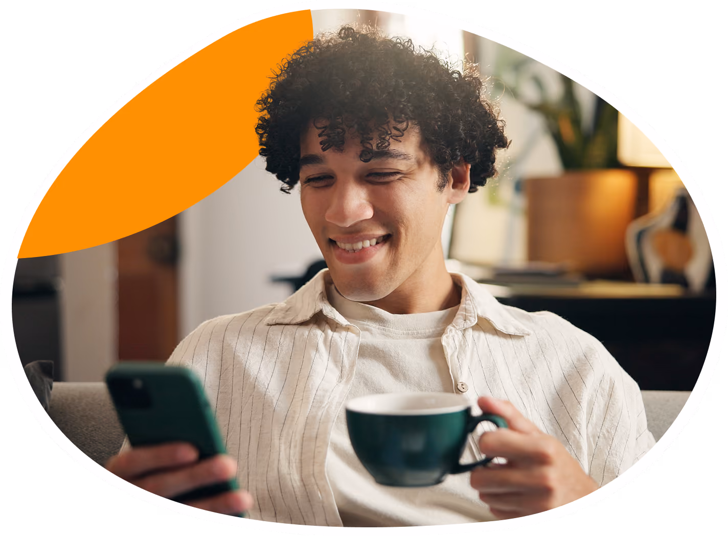 Person smiling while holding a smartphone in one hand and a green coffee cup and saucer in the other, sitting indoors on a couch