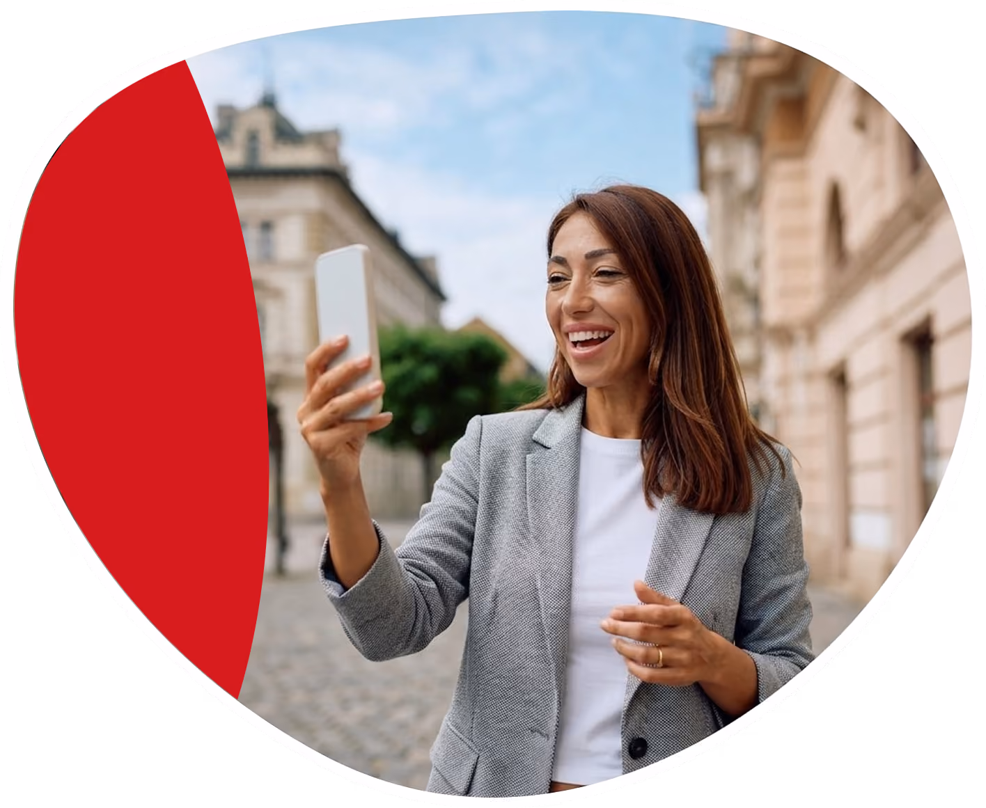 Smiling person holding a smartphone outdoors on a cobblestone street with buildings and trees in the background.