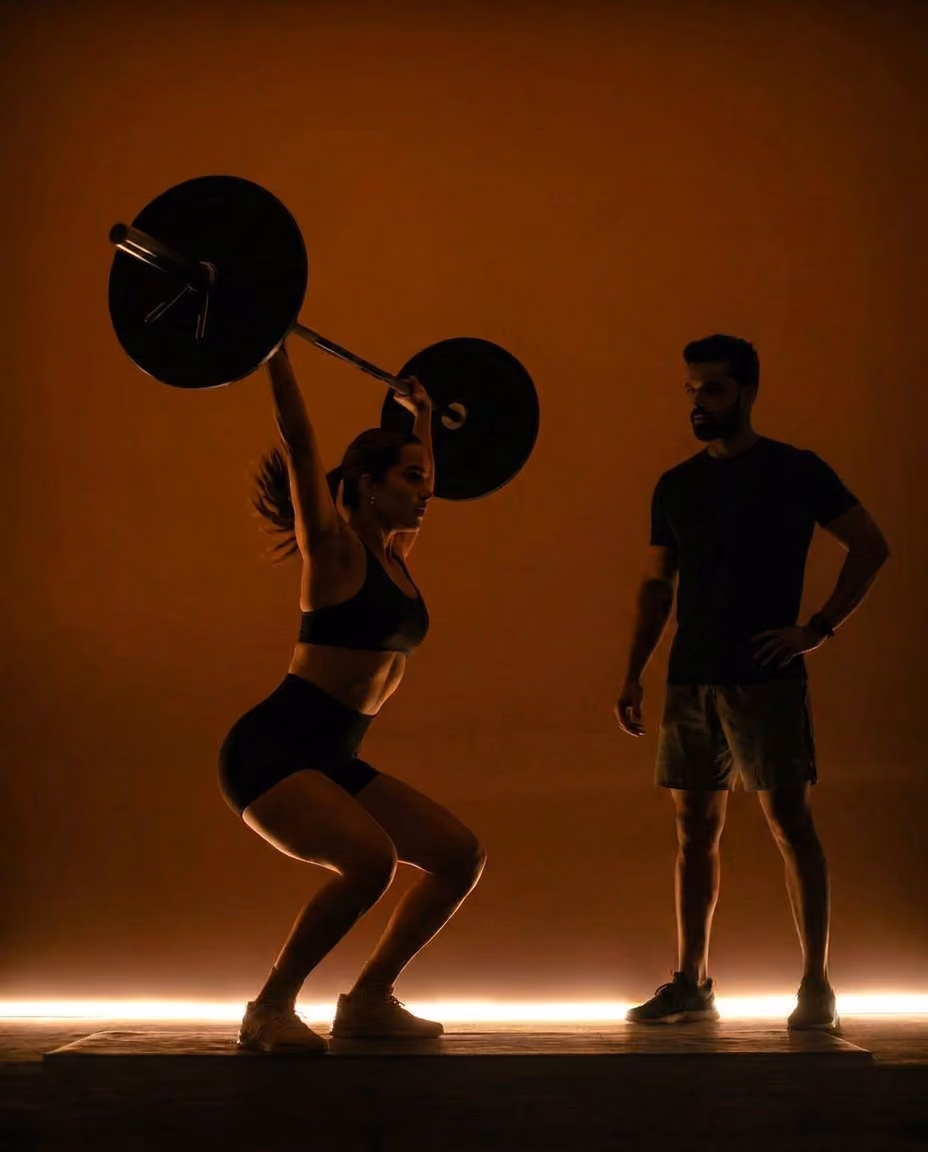Two people in a gym, one doing an exercise with a barbell, the other watching them giving them support