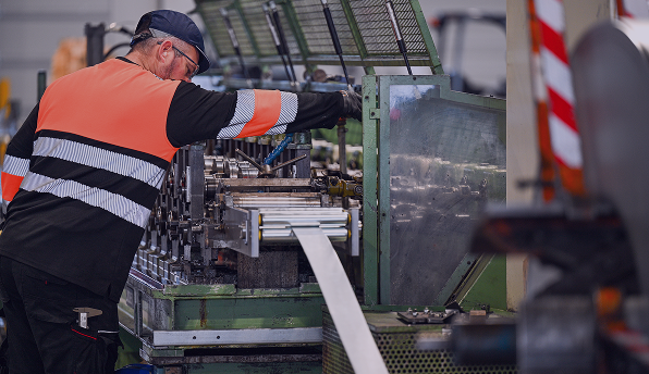 Un opérateur en tenue de travail orange et noire et casquette noire ajuste une machine industrielle fabriquant une bande métallique dans un atelier de production.