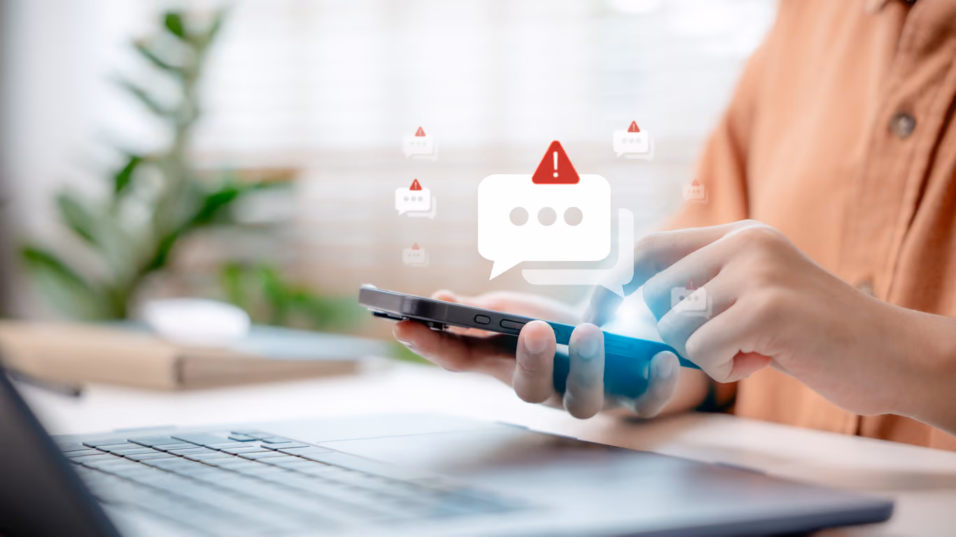 Eine Person hält ein Smartphone in der Hand und tippt darauf, umgeben von mehreren digitalen Warnsymbolen wie Ausrufezeichen in Sprechblasen, neben einem Laptop auf einem Schreibtisch.
