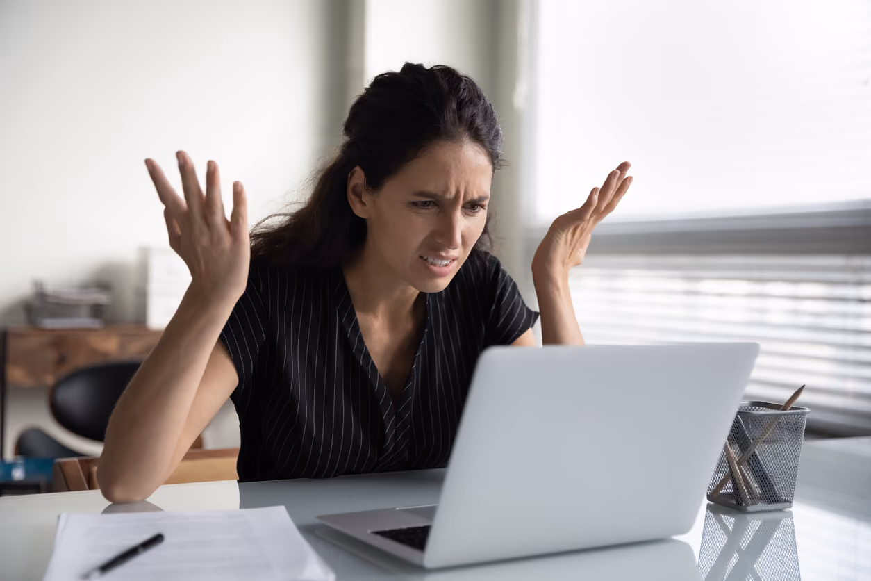 Eine Frau sitzt an einem Tisch vor einem Laptop und zeigt mit hochgehobenen Händen und einem verärgerten Gesichtsausdruck Frustration.