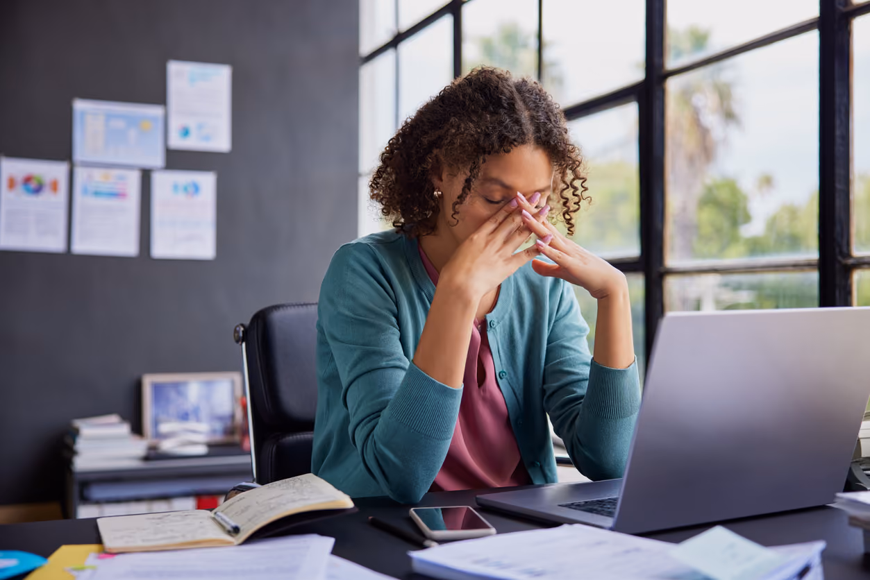 Eine Frau sitzt an einem Schreibtisch mit Laptop, Smartphone, offenem Notizbuch und Papieren und reibt sich müde das Gesicht in einem Büro mit großen Fenstern.