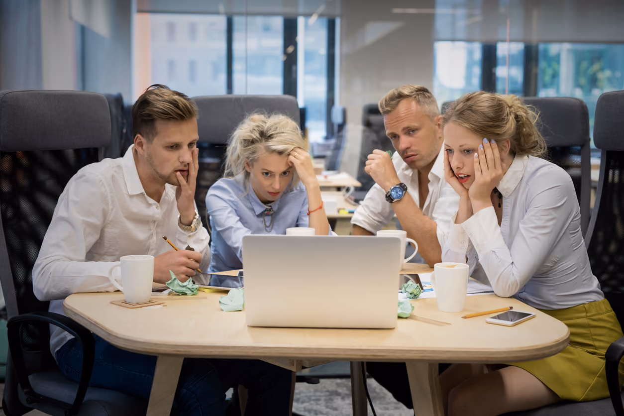 Vier junge Erwachsene sitzen in einem Büro um einen Tisch und schauen konzentriert auf einen Laptop, offenbar in einer intensiven Besprechung oder Problemlösung.