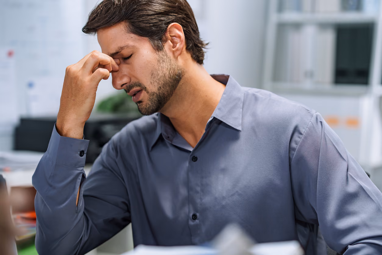 Ein junger Mann mit dunklem Haar und Bart sitzt in einem Büro und hält sich mit geschlossenen Augen den Nasenrücken, als ob er Kopfschmerzen oder Stress hätte.