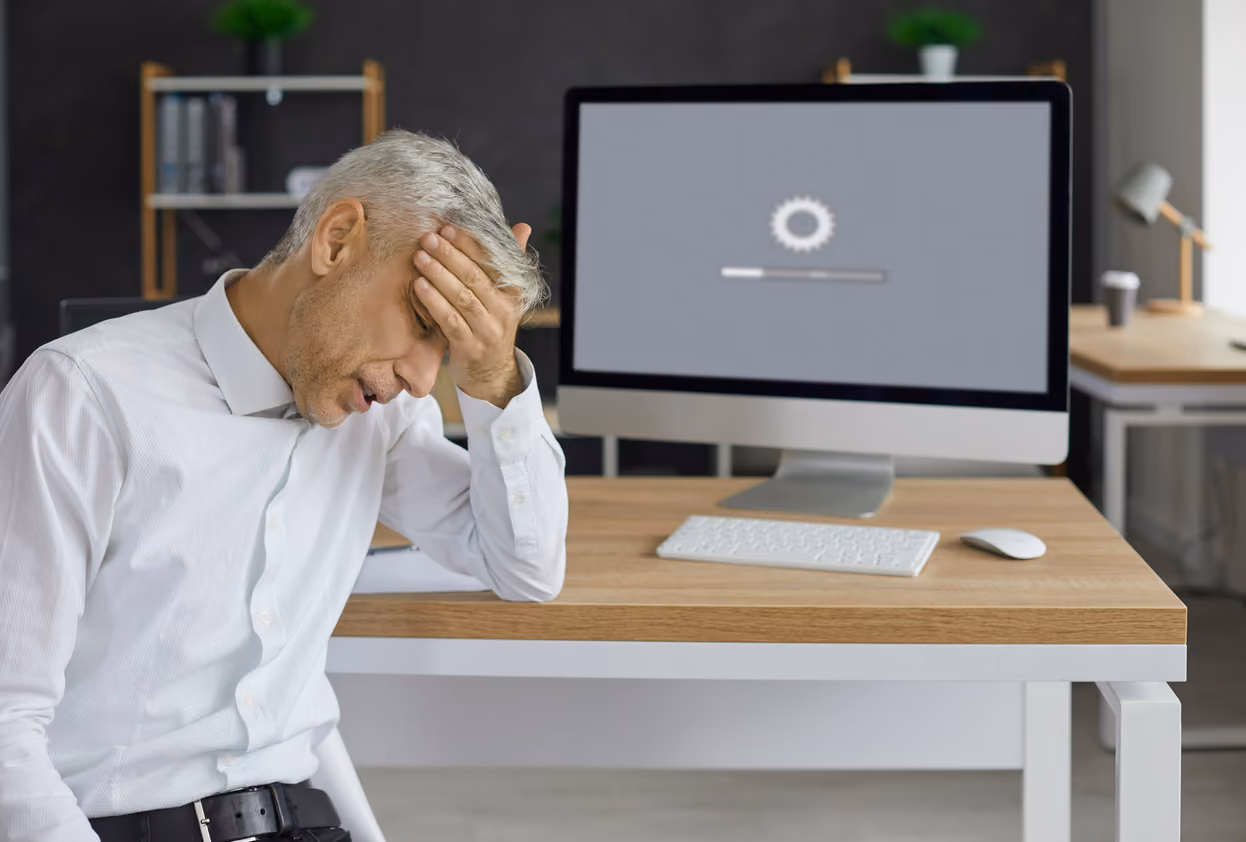 Älterer Mann mit grauen Haaren, der am Schreibtisch sitzt und sich den Kopf hält, während vor ihm ein Computer mit einem Ladebalken auf dem Bildschirm steht.