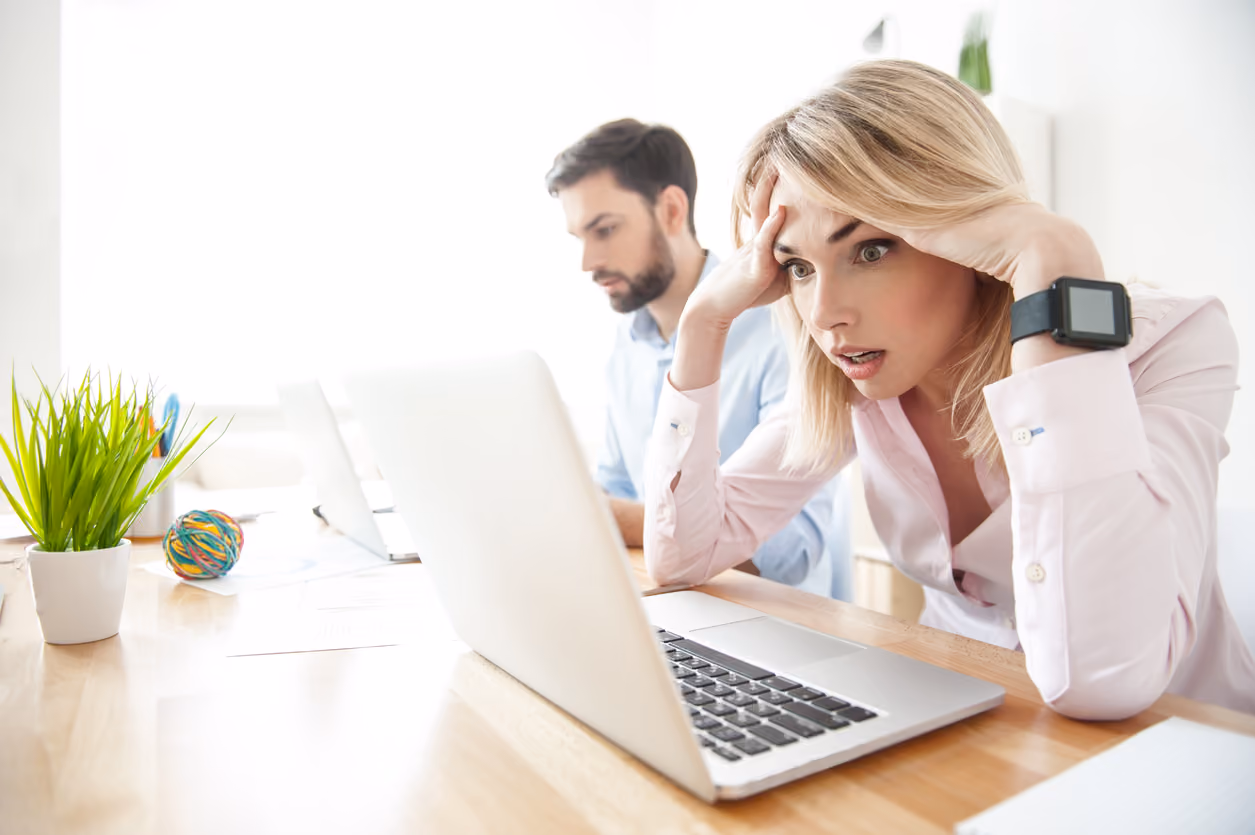 Eine Frau sitzt an einem Schreibtisch vor einem Laptop und sieht frustriert und besorgt aus, während ein Mann im Hintergrund an einem weiteren Laptop arbeitet.