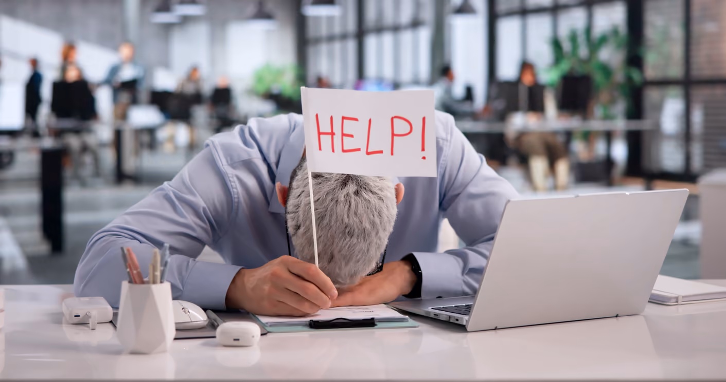 Ein Mann mit grauen Haaren sitzt an einem Schreibtisch mit einem Laptop und hat seinen Kopf auf den Tisch gesenkt, während er ein kleines Schild mit der Aufschrift ‚HILFE!‘ hält, im Hintergrund ein modernes Büro mit anderen Mitarbeitenden.