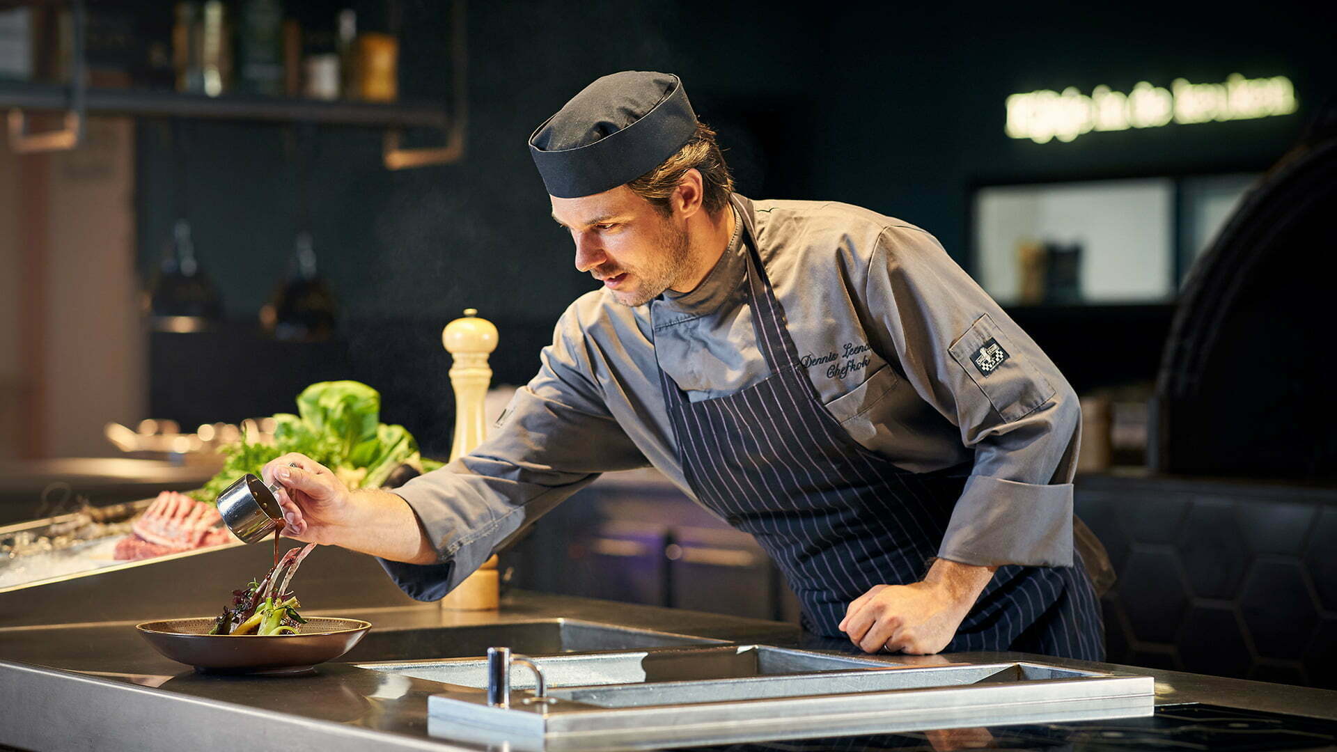 Chef in a grey chef's jacket plating a dish in an open kitchen