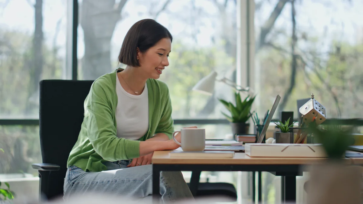 Woman in a green cardigan smiling while working on a laptop at a desk by a large window