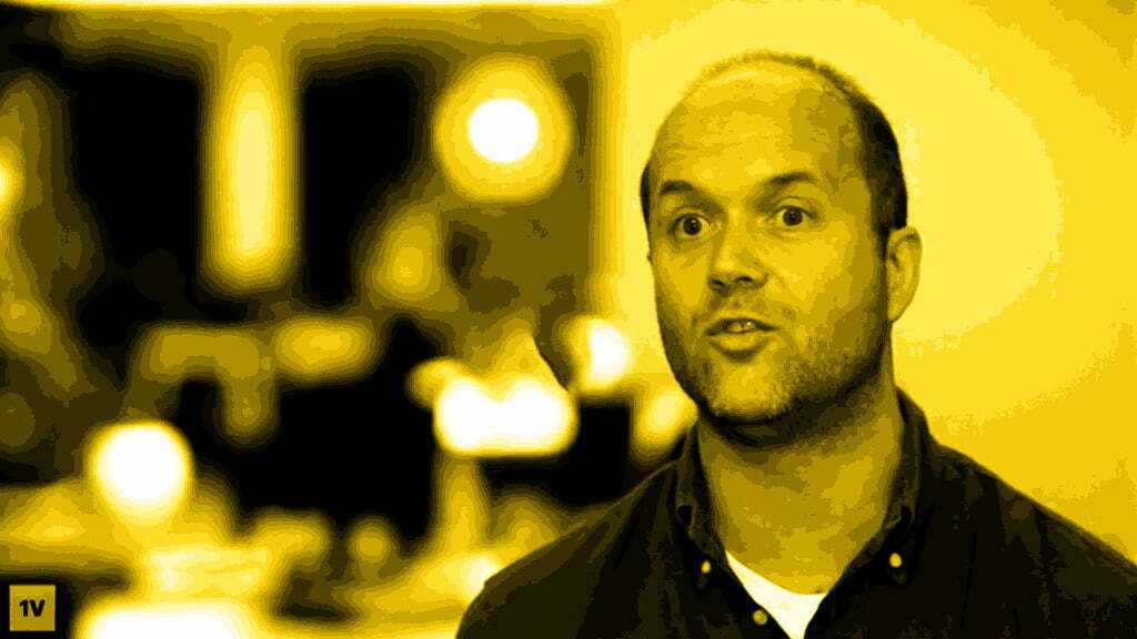 Bald man with a beard talking on camera, video still with a yellow colour filter
