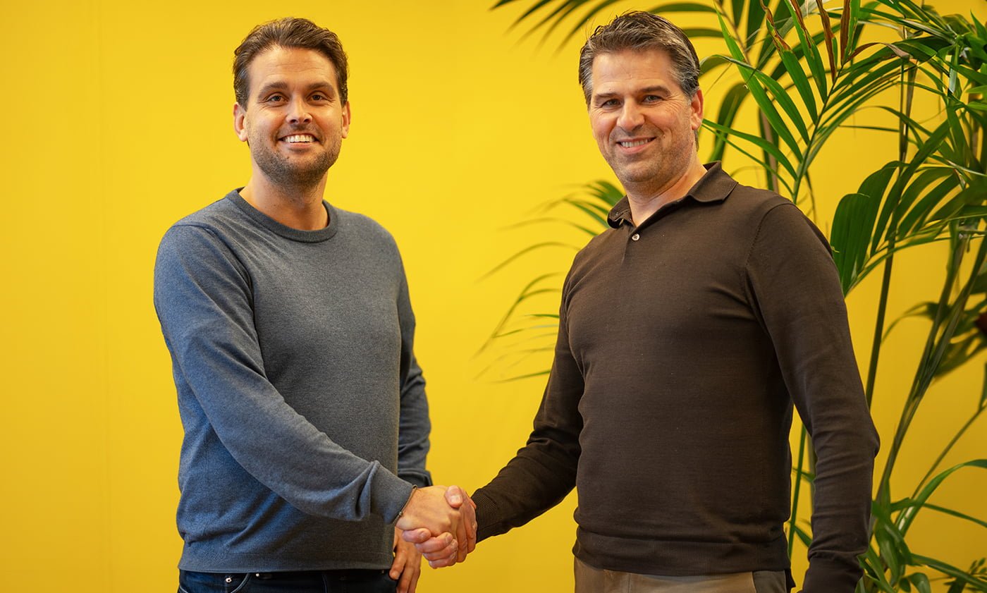 Two smiling men shaking hands in front of a yellow wall with a palm plant