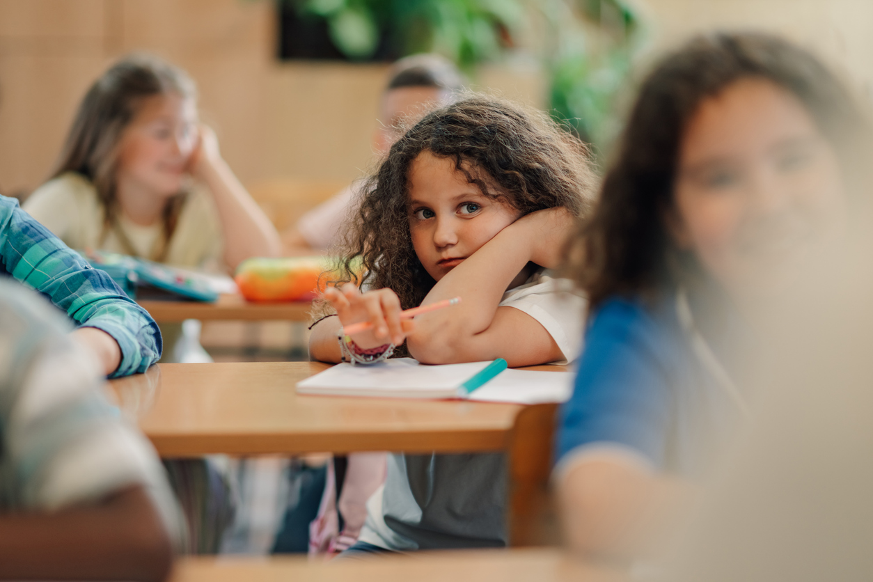 Girl with curly hair leaning on her hand in a classroom, looking at the camera with a pencil in her hand