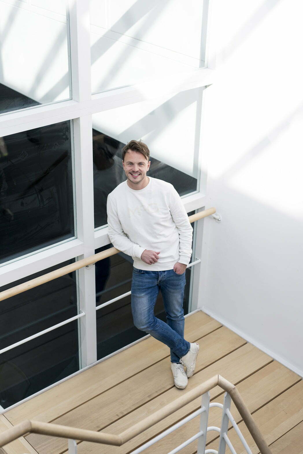 Smiling man in a white jumper and jeans leaning against a stair railing