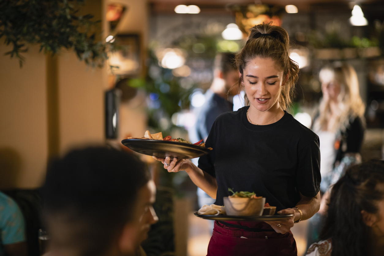 Waitress carrying two plates of food walks through a busy restaurant