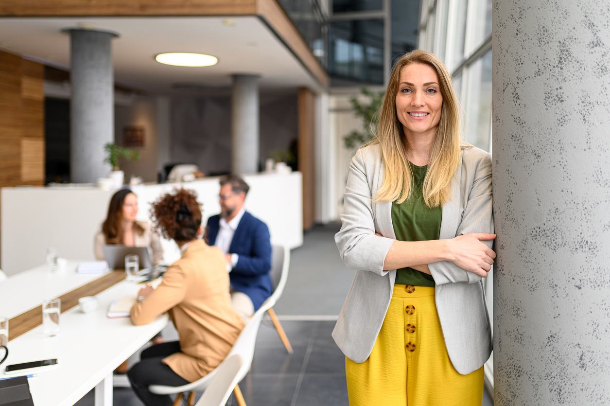 Smiling woman with folded arms leaning against a pillar, colleagues in a meeting in the background