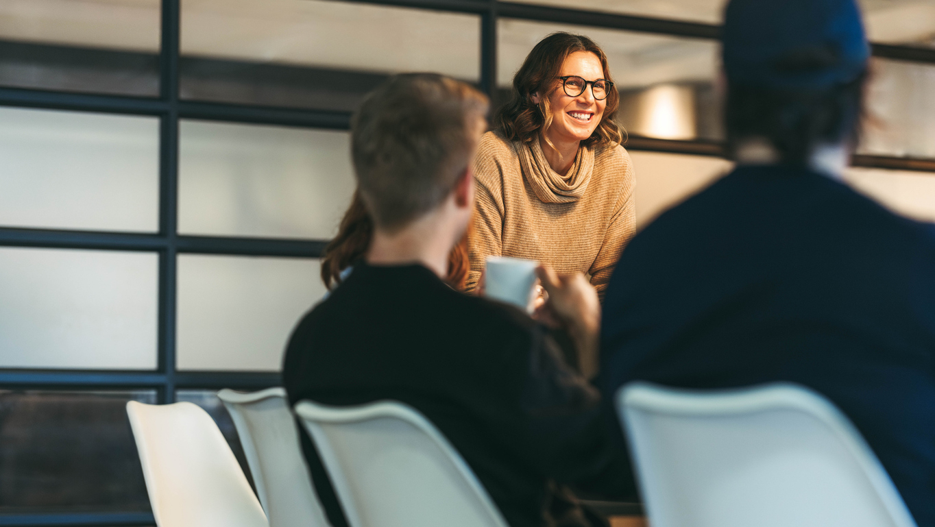 Smiling woman with glasses looking at colleagues in an office, with two people seen from behind in the foreground