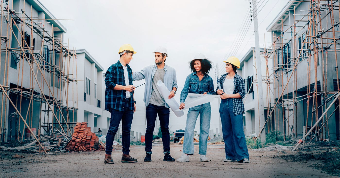 Four young construction workers in hard hats with building plans on a building site with scaffolding and houses