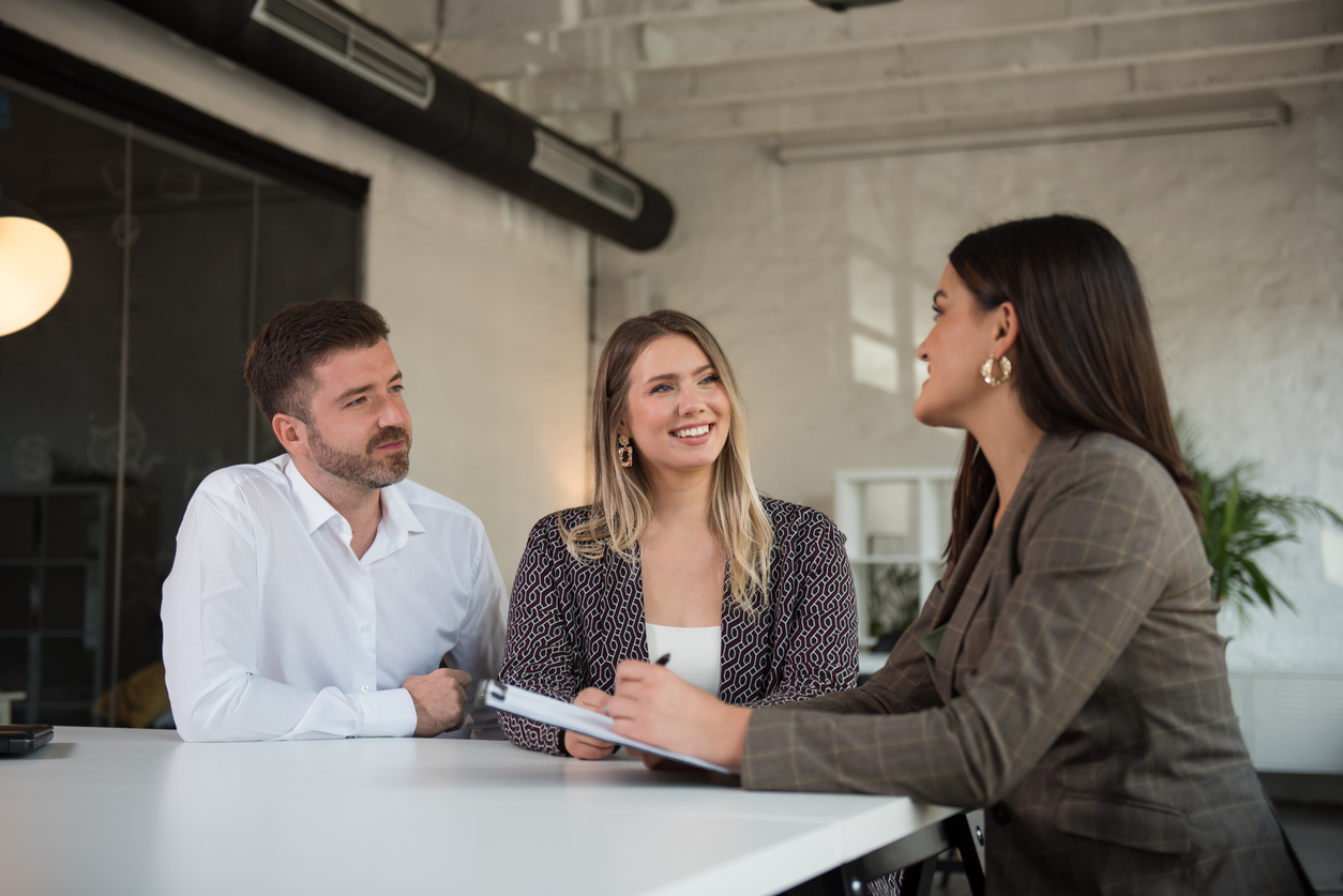 Three people talking at a white table, a woman writing something on a clipboard